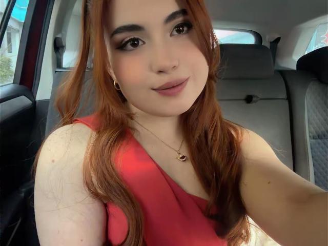VicluX, met rood haar, draagt een rode jurk, waarbij haar borsten zichtbaar zijn, zit in een auto, glimlacht, heeft make-up met donkere eyeliner, een ketting met een rode steen en houdt een telefoon vast..