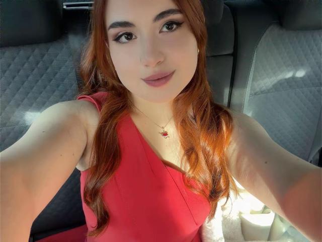 VicluX, avec ses cheveux roux, portant une robe rouge, a de petites seins et un visage mignon, elle prend un selfie dans une voiture, souriant avec un maquillage léger et un collier..