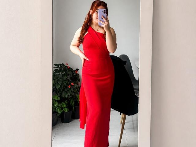 VicluX con vestido rojo, con un solo hombro, abertura lateral larga, sosteniendo un teléfono, cubriéndose los senos con la mano, selfie frente al espejo, cabello rojo, pared blanca, silla negra, planta verde, suelo blanco..
