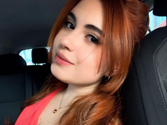 VicluX ha i capelli rossi, gli occhi marroni e una carnagione chiara, indossa una maglietta rosa, guarda la fotocamera con un leggero sorriso, seduta in un'auto..