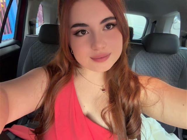 VicluX met lang rood haar, gekleed in een rode jurk, die haar borsten laat zien, zittend in een auto, lachend naar de camera, met een kleine ketting, lichte huid en subtiele make-up..