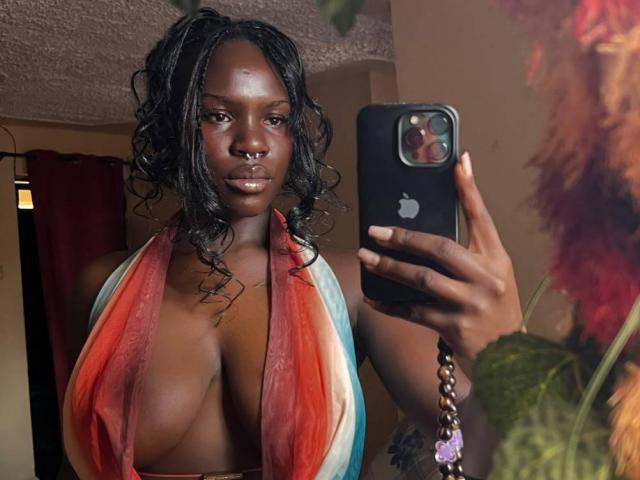 EmpressWater with dark skin, curly black hair, and large, shiny tits in a rainbow-colored, deep cleavage halter top, taking a selfie with a black iPhone in a dim room.