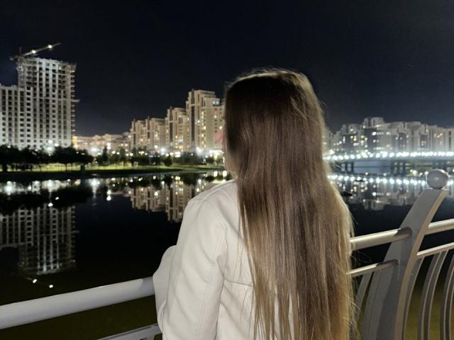 LeanaBairos with long brown hair, white shirt, standing on a bridge at night, facing a city's reflection in a calm waterway, city lights illuminated, urban skyline in background.
