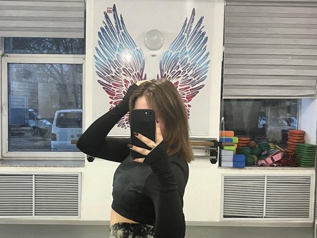 LeanaBairos, con un busto de tamaño medio, vestida con una camiseta negra de manga larga y pantalones de camuflaje, se está tomando un selfie frente al espejo en un gimnasio, cubriendo su rostro con un teléfono..