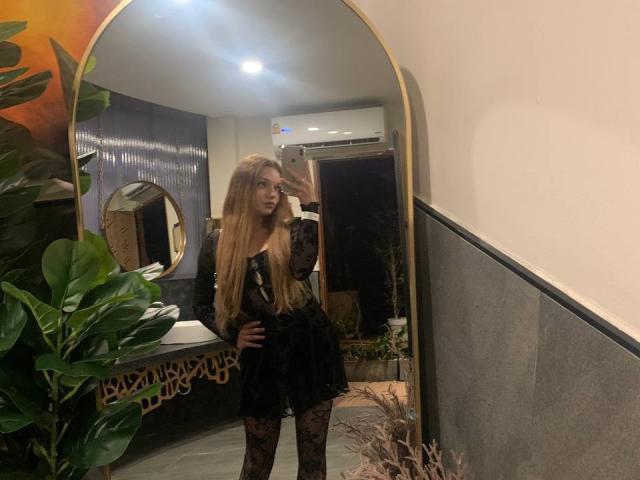 LeanaBairos, met lang blond haar, een zwarte kanten jurk en kanten leggings, staat voor een spiegel en maakt een selfie, waarbij haar achterkant te zien is, in een kamer met weinig licht..