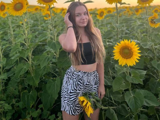 LeanaBairos met lang bruin haar, een zwarte top zonder bandjes, een rok met een zebraprint, staand in een veld met zonnebloemen, een bloem vasthoudend, hand in haar haar, lachend, zonsondergang op de achtergrond..