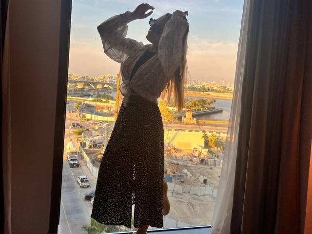 LeanaBairos with long hair, back to camera, wearing sheer top and black dotted skirt, raising arm, standing in front of window, sunlight on her ass, city view outside.