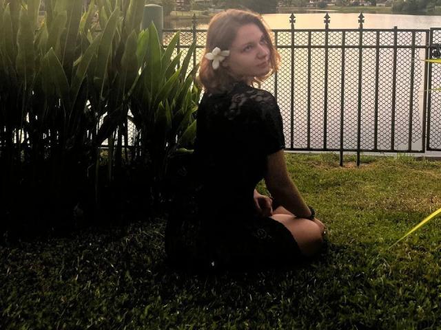 LeanaBairos, con una flor blanca en su cabello castaño, está arrodillada sobre la hierba, vestida con un vestido negro de encaje, mirando por encima del hombro, con una valla de metal y agua al fondo..
