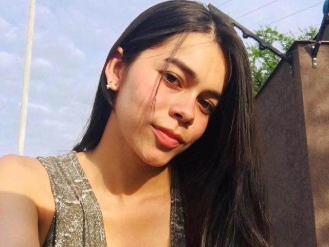 AlanaBlake tem cabelos pretos longos, pele clara, seios pequenos e um rosto bonito com um sorriso discreto, vestindo uma blusa cinza sem mangas, ao ar livre sob a luz forte do sol..