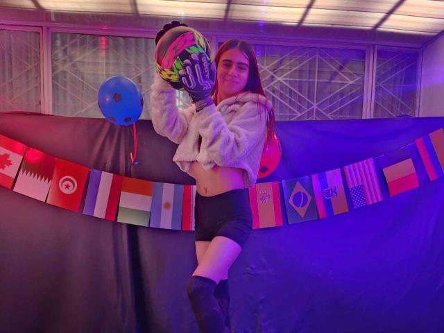 LoveMaxim with long brown hair, wearing a fluffy white crop top, black shorts, and gloves, holding a helmet, standing with a purple-blue glow, and colorful flags in the background.