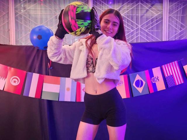LoveMaxim with long brown hair, smiling, holding a colorful helmet, wearing a fluffy white jacket, black shorts, and black gloves, has a belly button piercing, and cute tits.