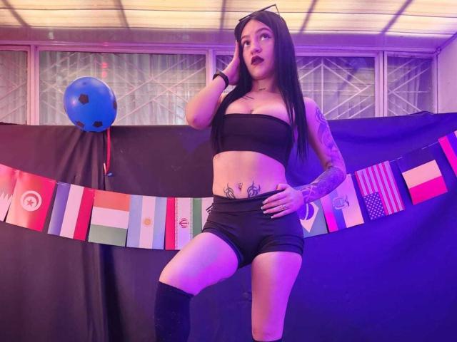 ViolettaSquirt, con cabello largo y negro, vestida con un top negro ajustado, pantalones cortos negros y calcetines hasta la rodilla, de pie con una mano en la cadera, mostrando tatuajes e iluminación morada-rosa..