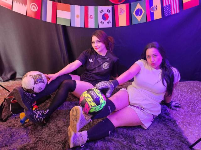 SaraAndMarilyn69 sedute su un tappeto peloso, vestite con abbigliamento da calcio, con magliette bianche e nere, calze nere fino al ginocchio e palloni da calcio, in una stanza illuminata di viola, sorridono alla telecamera..