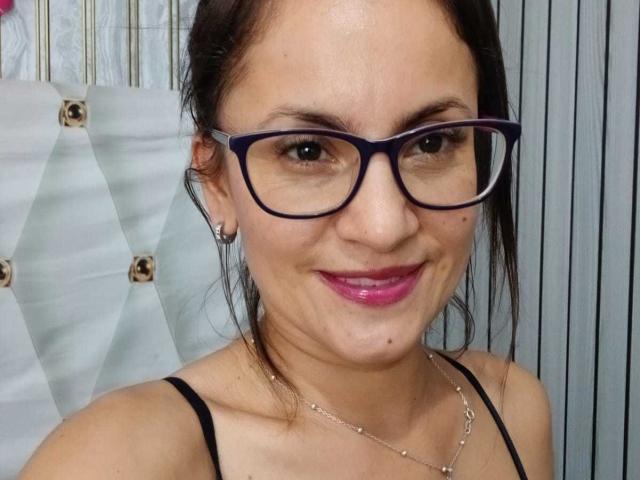 SolaraDubois with big blue glasses, pink lipstick, and a smile, wearing a black spaghetti strap top, showing a cute face and small tits.