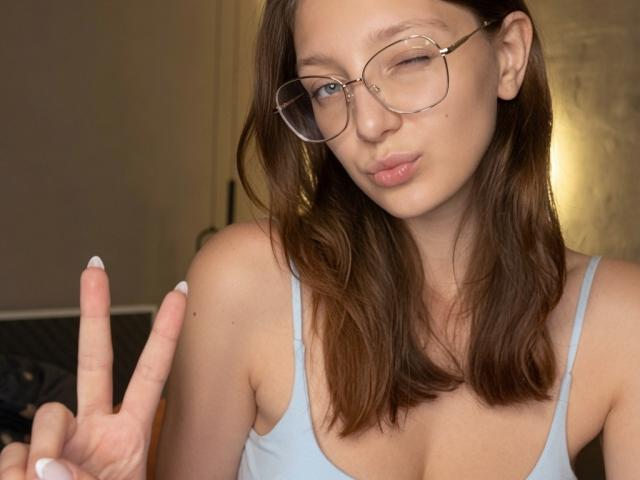 Stacey Averbach, con gli occhiali, che fa l'occhiolino e mostra il segno della pace, indossa una canotta azzurra, che mette in mostra il suo seno piccolo e il suo viso carino, con i capelli castani e le unghie bianche..
