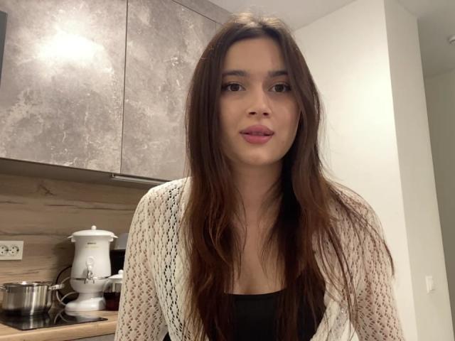 LylaFinne, con cabello castaño largo, labios rosados, vestida con un cárdigan blanco de punto sobre una blusa negra, está de pie en una cocina con un armario gris y electrodomésticos blancos..