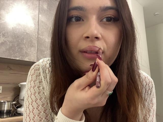 LylaFinne, com longos cabelos castanhos, vestindo uma blusa branca de renda, chupa um batom vermelho com suas unhas pintadas de vermelho, olhando para a câmera com a boca ligeiramente aberta..