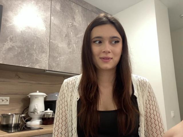 LylaFinne has long brown hair, a small nose, and is wearing a white patterned cardigan over a black top. She's looking to the side with a slight smile.