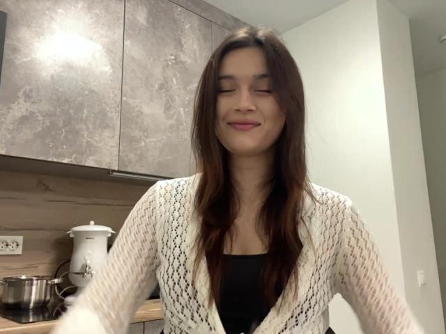 LylaFinne, con lunghi capelli castani, sorride con gli occhi chiusi, indossa un cardigan bianco in maglia, una maglietta nera e rossetto rosa, ed è in piedi in una cucina moderna..