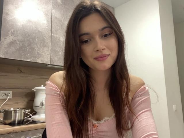 LylaFinne, con lunghi capelli castani, una maglietta rosa a spalle scoperte e un trucco leggero, sorride dolcemente in una cucina. Il suo seno è piccolo e la sua vulva non è visibile..