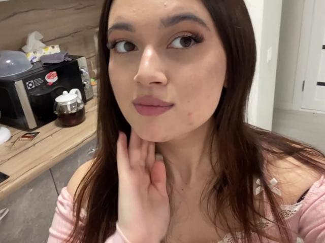 LylaFinne mit braunen Haaren, heller Haut und rosafarbenem Lippenstift, die sich ihr Kinn berührt, trägt ein rosa Top mit einem Schulterauschnitt, hat eine kleine Pickel auf ihrer Wange und hat braune Augen..