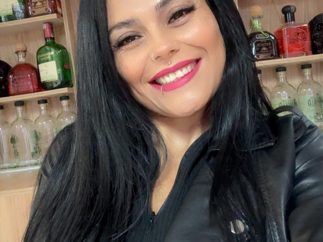 CharlotteCrofort with long black hair, smiling, pink lipstick, black leather jacket, nose ring, and visible breasts in a bar setting with bottles in the background.