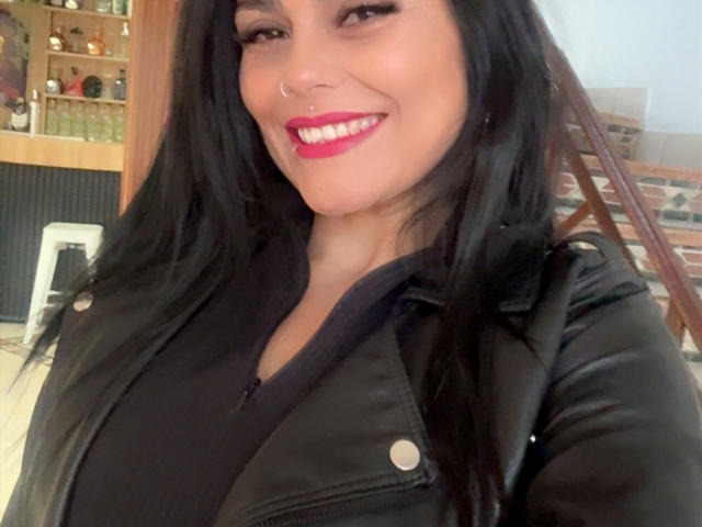 CharlotteCrofort with black hair, nose ring, and pink lipstick, smiling, wearing a black leather jacket, showcasing her large tits, in a selfie.