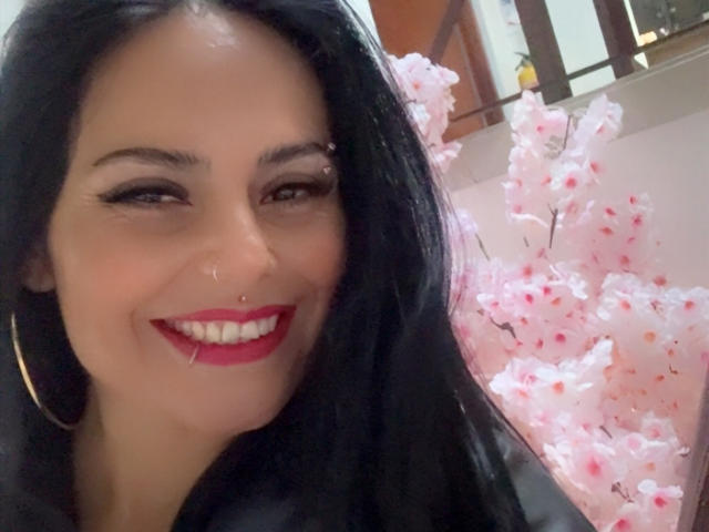 Charlotte Crofort, com cabelo preto, batom vermelho, brincos de argola, piercing no nariz, sorrindo, mostrando os dentes brancos, flores de cerejeira rosa ao fundo, luz suave, ligeiramente desfocada..
