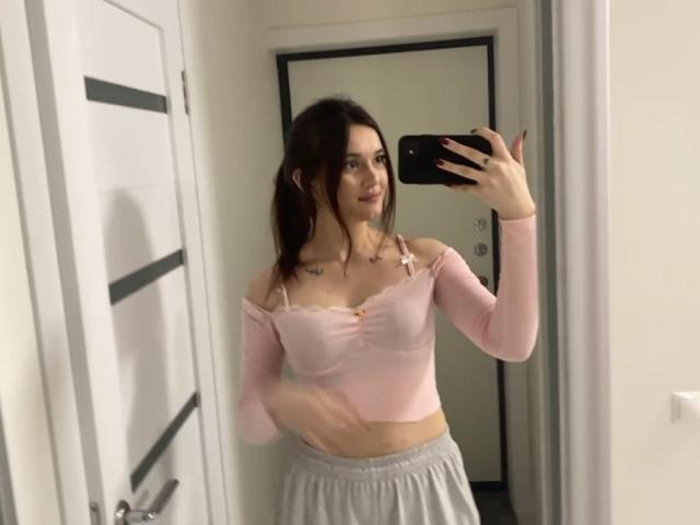 LylaFinne si scatta un selfie allo specchio, indossando una maglietta rosa con una spalla scoperta che mette in mostra i suoi piccoli seni, e pantaloni da jogging grigi, con tatuaggi sul petto, mentre tiene in mano un telefono nero..