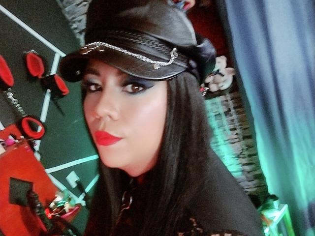 LexyDom, com cabelo preto, batom vermelho e boné de couro preto, olhando para a câmera, vestindo uma blusa preta, em um quarto com temática BDSM, com brinquedos sexuais vermelhos e pretos..