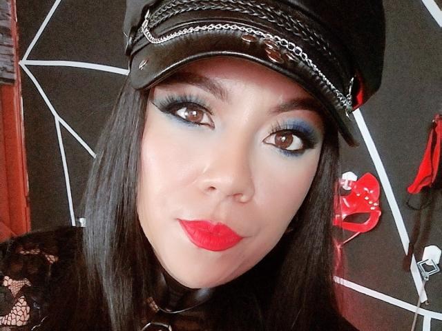 LexyDom mit schwarzer Lederkappe, blauem Lidschatten, rotem Lippenstift und langen schwarzen Haaren. Ihre braunen Augen sind halb geöffnet, was ihr einen verführerischen Blick verleiht. Sie trägt eine schwarze Spitzenbluse..