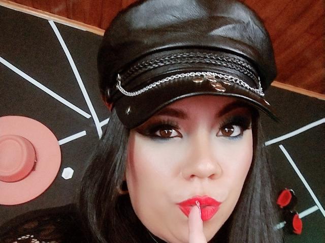 LexyDom tem cabelo escuro, usa um boné de couro preto e batom vermelho, com o dedo tocando os lábios, exibindo uma maquiagem de olhos marcante e uma expressão levemente emburrada..