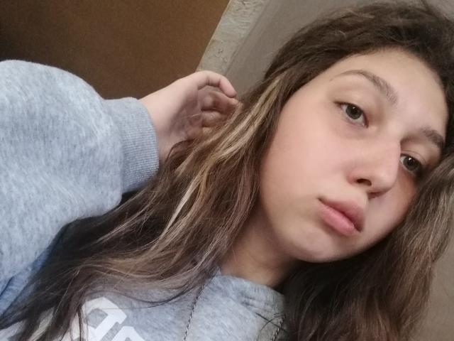 Sanella with pouty lips, long brown hair, and a gray hoodie, looking at the camera with a neutral expression. Her hand is in her hair.