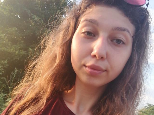 Sanella's face is in a selfie, with wavy brown hair, subtle makeup, and a slight smile. Her small nose and full lips are visible. She's wearing a red shirt.