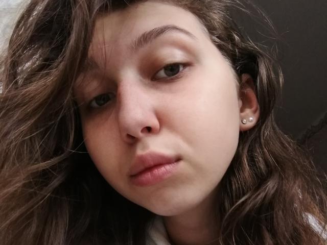 Sanella has wavy brown hair, fair skin, and small earrings. She has green eyes, full pink lips, and a slightly serious expression. Her nose is small and she's looking forward.
