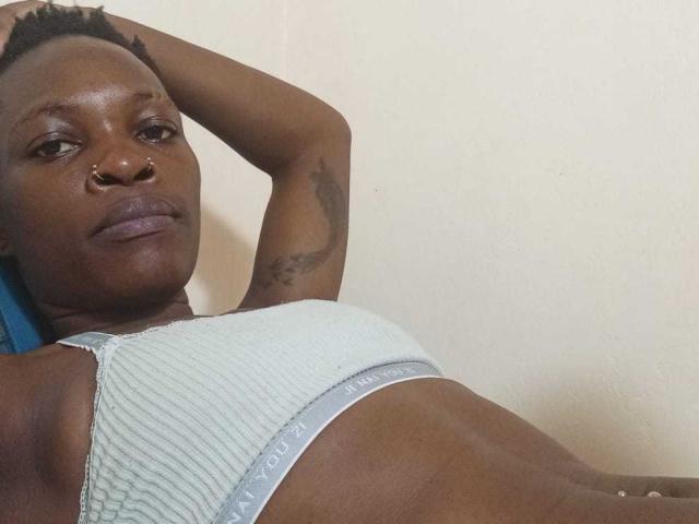 MelanyPrincess has dark skin, short black hair, nose ring, tattoo on arm, wearing a gray Calvin Klein bra, lying down with arm behind head, looking at camera.