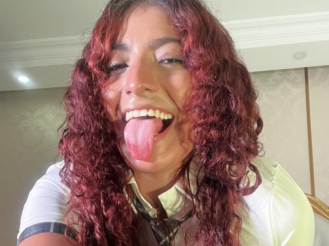 Mia Bianchi, con i capelli rossi ricci, sorride con gli occhi chiusi e la lingua fuori, indossa una camicia bianca con bordi neri e una cravatta a fantasia, appare carina e giocosa..