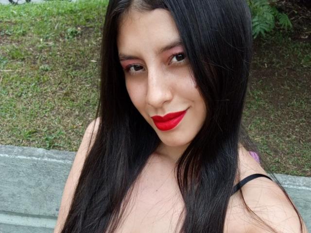 MadissonFontaine with long black hair, red lipstick, pink eyeshadow, and black strap dress, smiling at the camera, showing her tits slightly. Her ass is not visible.