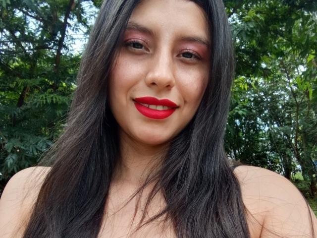 MadissonFontaine with long dark hair, red lipstick, and pink eyeshadow, smiling at the camera, standing in front of green trees. Boobs not visible.