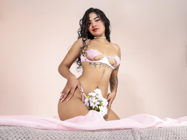ZoeClarkk, con cabello negro rizado, sostén y tanga rosas, sosteniendo flores blancas, arrodillada sobre tela rosa, mostrando sus pechos y vulva, tatuaje en el lado derecho, collar con un lazo al cuello..
