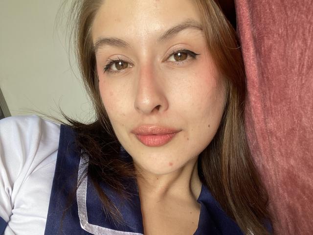 Isabella Palacio hat helle Haut, braune Augen und braunes Haar. Sie trägt ein blau-weißes Hemd, hat eine kleine Pickel auf ihrem Kinn und rosafarbene Lippen..