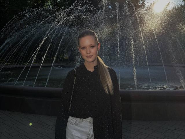 Lilliana Whang indossa una camicia nera a pois, pantaloni bianchi a vita alta e ha una lunga coda di cavallo. È in piedi di fronte a una fontana d'acqua con getti illuminati dalla luce del sole..