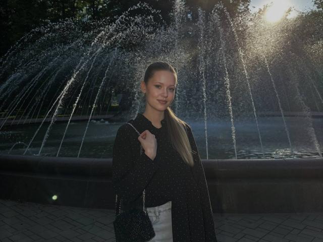 Lilliana Whang steht vor einem Brunnen, trägt ein schwarzes, gepunktetes Hemd und weiße Hosen, hat eine schwarze Tasche bei sich und lächelt. Ihr langes, braunes Pferdchen fängt das Sonnenlicht ein..