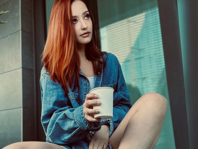 De aspecto atractivo y con el cabello rojo, sentada con las piernas cruzadas, vistiendo una chaqueta vaquera azul, sosteniendo una taza blanca, mirando hacia un lado, con una expresión linda, relajada y sensual..