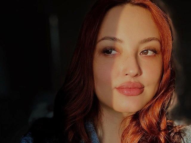 Cherryy-hot tem cabelo ruivo, pele clara e lábios rosados e cheios. A luz do sol projeta uma sombra em seu rosto. Ela está olhando para o lado com uma expressão levemente emburrada..