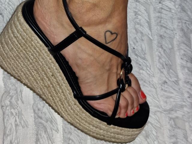 Il piede di Cassandra Michelli in un sandalo nero con zeppa e suola in paglia intrecciata, che mostra un piccolo tatuaggio a forma di cuore sopra la caviglia e le unghie dei piedi dipinte di rosso..