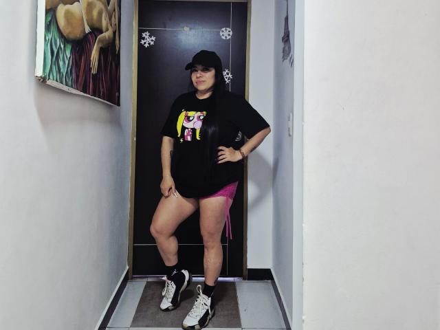 EvaLunaLatina está em um corredor, vestindo uma camiseta preta, shorts rosa, tênis brancos, meias pretas e um boné preto, com a mão no quadril, exibindo suas nádegas..