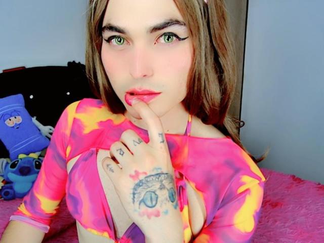 Anahi: Grüne Augen, Oberteil in Rosa und Gelb im Batik-Stil, Finger an der Unterlippe, tätowierte Hand, rosa Bett, braunes Haar und roter Lippenstift. Brüste sind durch das Oberteil sichtbar..