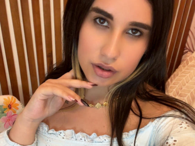 KatieDesirX with dark brown hair, light brown eyes, and pink lips, wearing a white off-shoulder top, touching her neck with a gold necklace, looking at the camera.