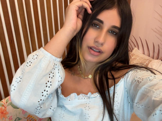 KatieDesirX tem cabelos longos e pretos com mechas loiras, vestindo uma blusa azul clara com um ombro de fora e mangas bufantes, olhando para a câmera com um leve sorriso..